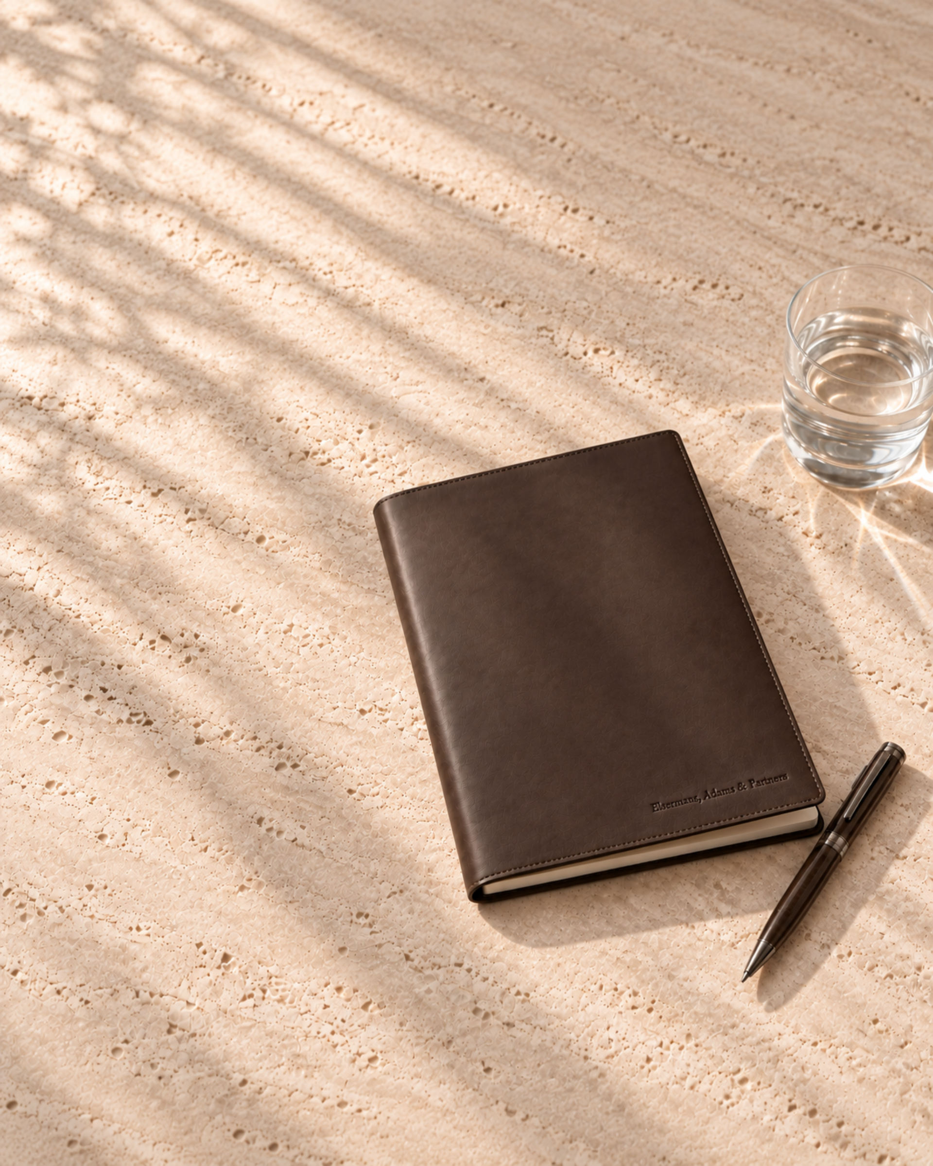 Leather notebook, pen and a glass of water on a travertine table in palm shade